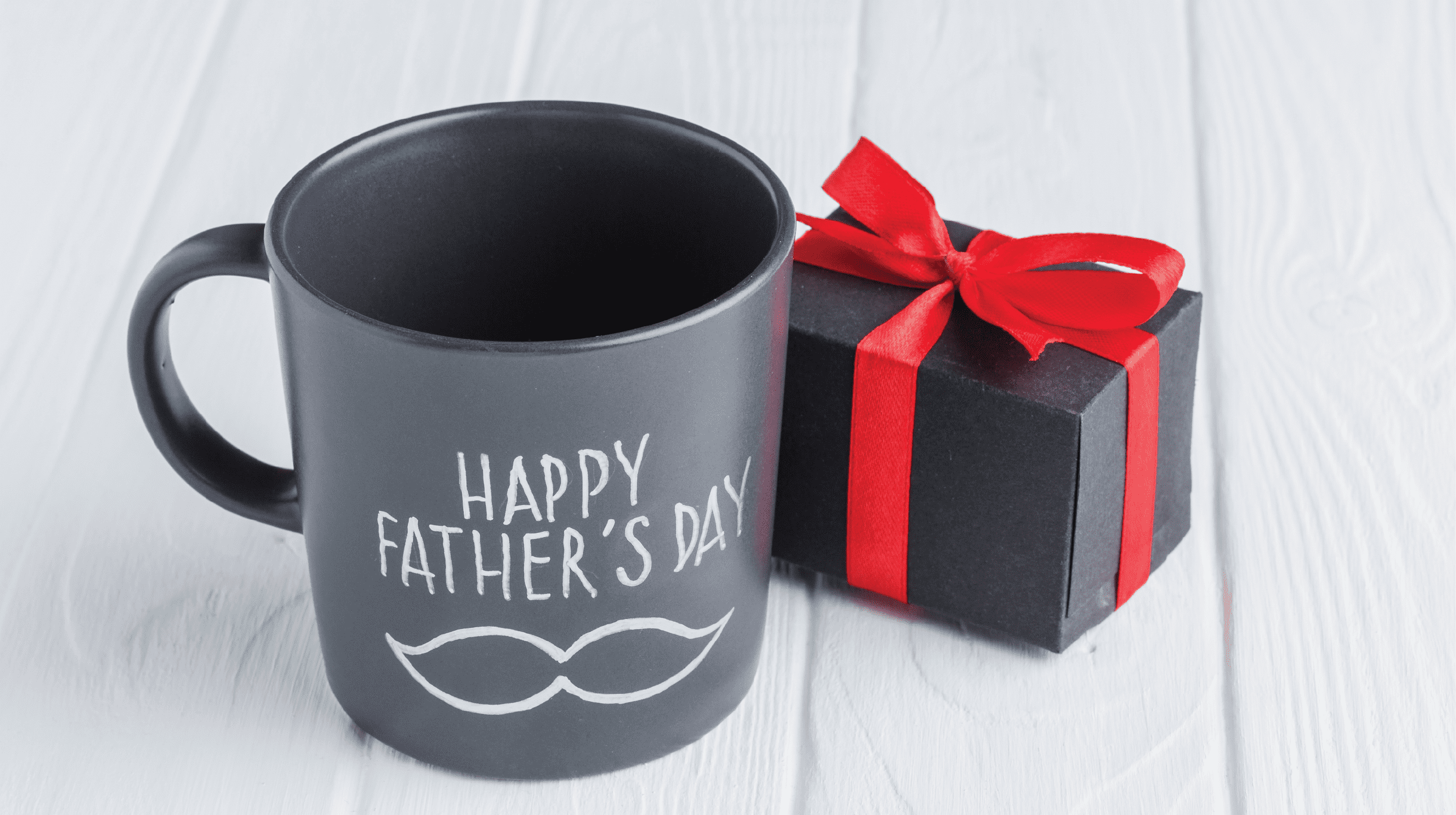 "Custom printed matte black ceramic mug with 'HAPPY FATHER'S DAY' text and a mustache illustration, placed next to a black gift box tied with a red satin ribbon on a white wooden surface."