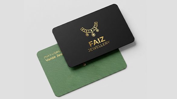 "An elegant top view of two rounded-corner business cards stacked together, featuring a matte black top card with a shiny gold foil FAIZ JEWELLERY logo and a textured forest green bottom card with gold Arabic and English typography."
