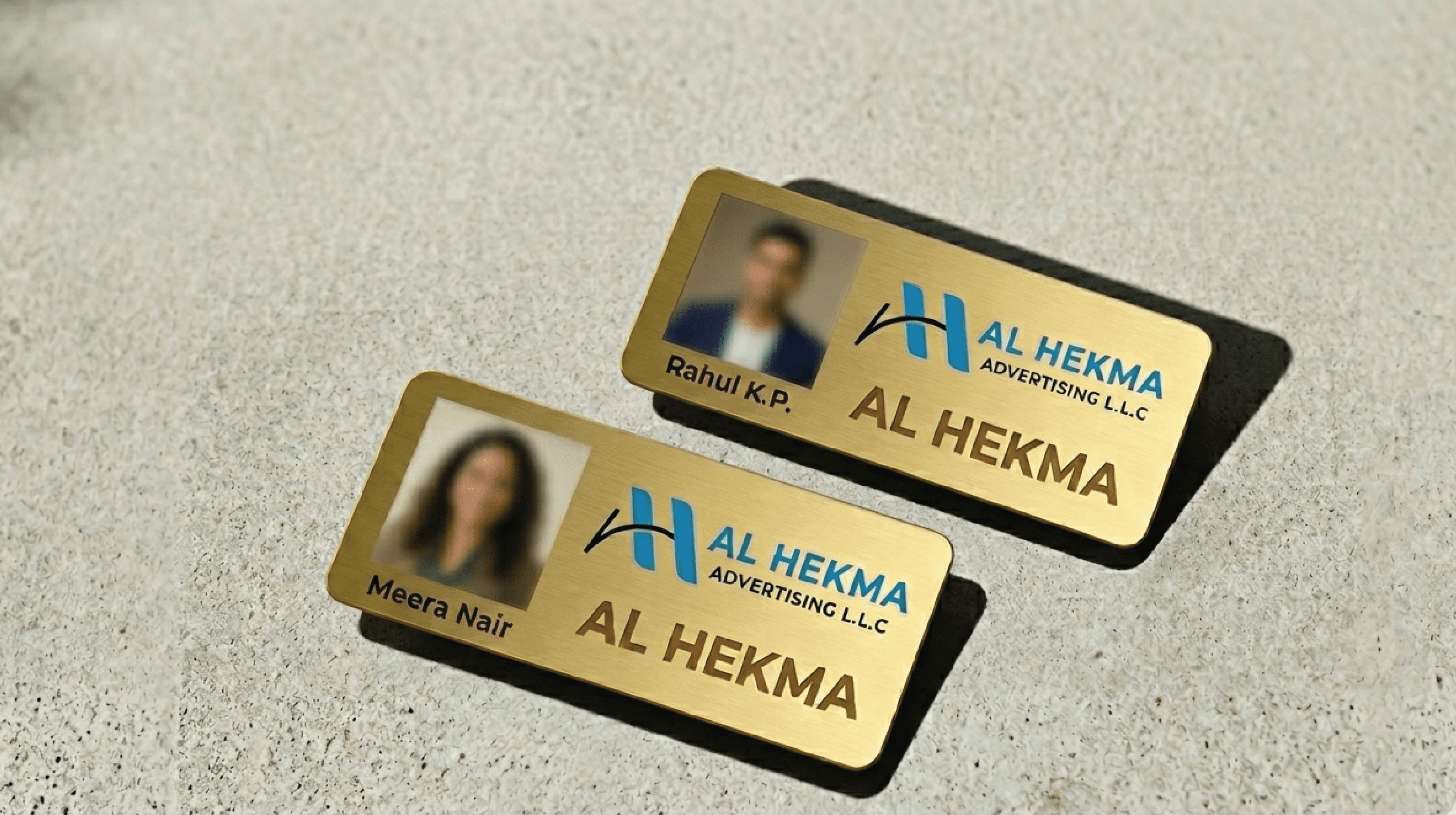 "Premium custom gold metal staff name badges for Al Hekma Advertising L.L.C featuring employee photos, names like Rahul K.P. and Meira Nair, and the company logo."