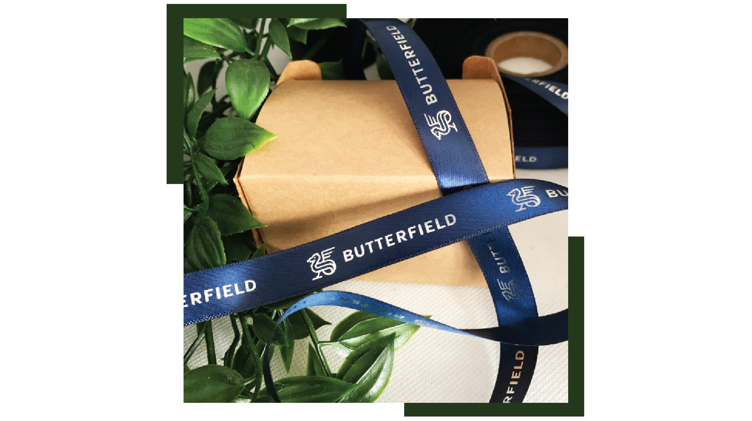 "Premium dark blue satin ribbon custom printed with the 'BUTTERFIELD' brand name and a dragon logo, wrapped around a small brown kraft gift box next to green leaves."